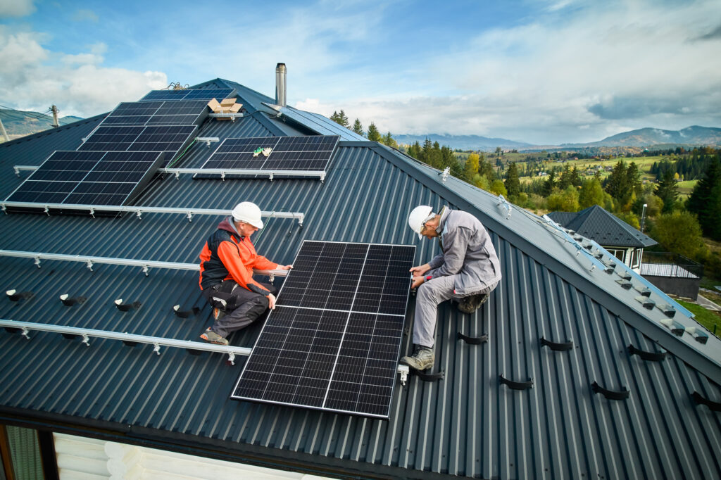 Zwei Handwerker mit weißen Schutzhelmen installieren ein Solarmodul auf einem steilen, dunkelgrauen Blechdach vor einer ländlichen Berglandschaft.