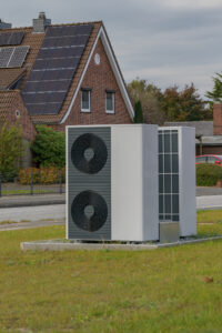 Eine moderne Luft-Wasser-Wärmepumpe steht vor einem Gebäude mit Photovoltaik auf dem Dach. Diese Kombination macht eine neue Wärmepumpe im Mehrfamilienhaus wirtschaftlich rentabel.