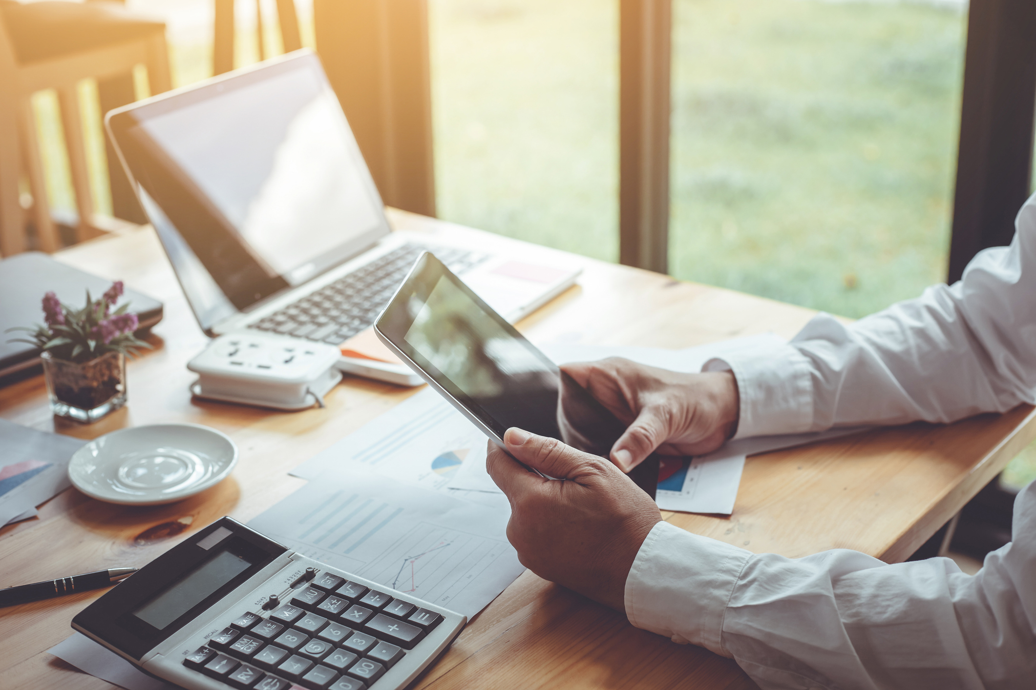 Eine Person im weißen Hemd sitzt an einem hellen Holztisch und hält ein Tablet in den Händen. Auf dem Schreibtisch befinden sich ein geöffneter Laptop, ein Taschenrechner, verschiedene gedruckte Diagramme, ein Stift sowie eine kleine Zimmerpflanze und eine Kaffeetasse. Im Hintergrund ist durch ein Fenster ein Garten erkennbar.