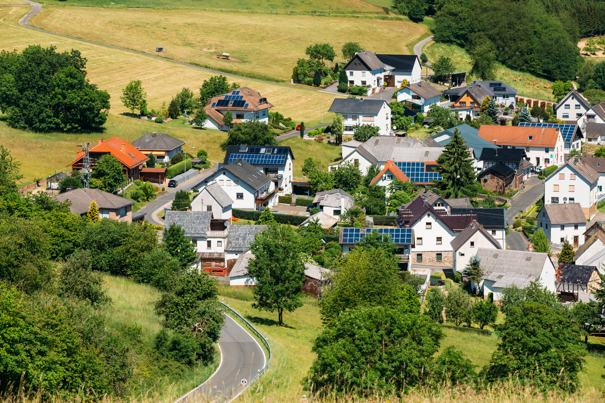 Eine Luftaufnahme eines ländlichen deutschen Dorfes in einer hügeligen Landschaft. Zahlreiche Wohnhäuser haben Photovoltaik-Solarmodule auf ihren Dächern, was das Vorbild der Kommunen für lokale Solarstromproduktion und nachhaltige Zusatzeinnahmen als Vermieter demonstriert.