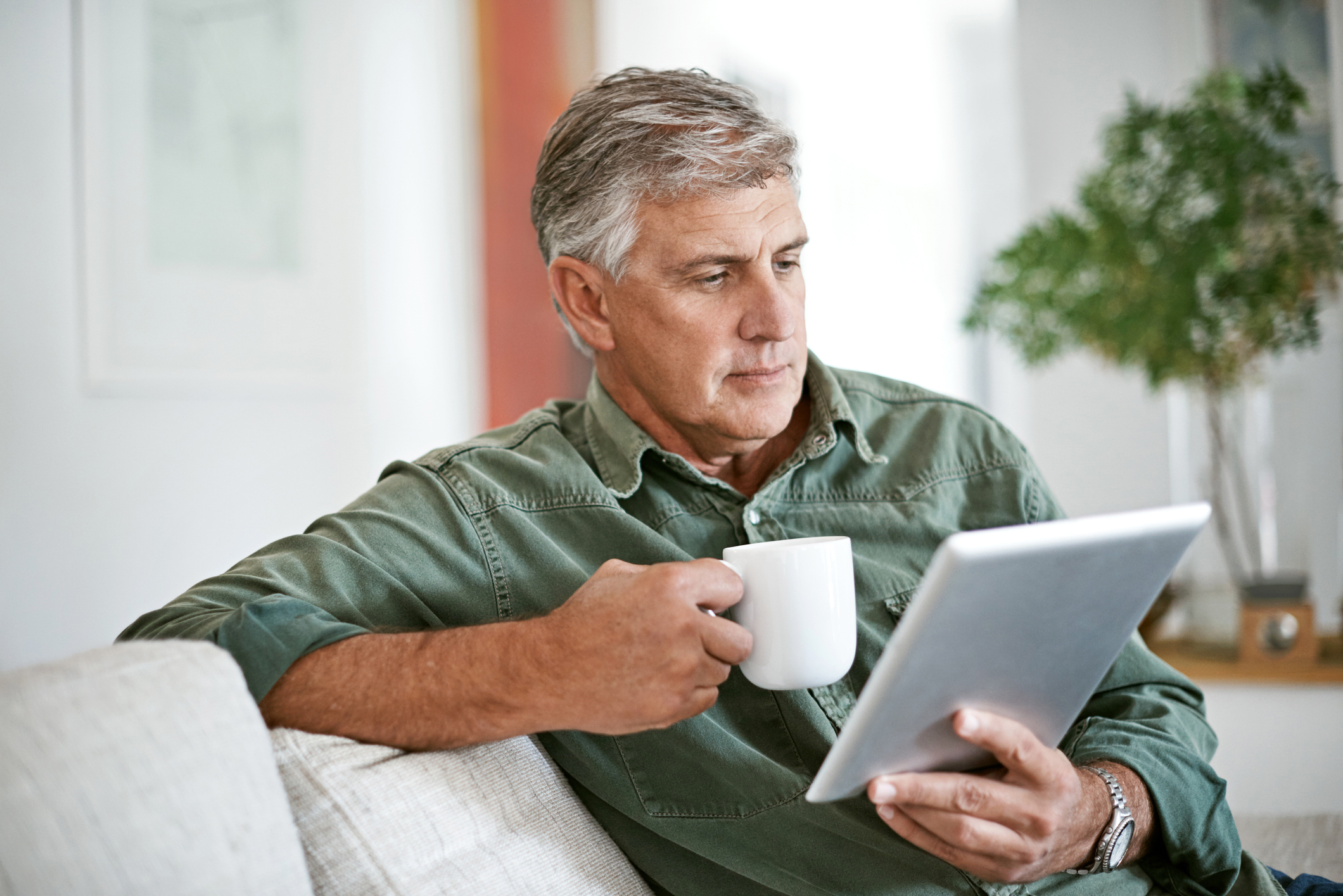 Ein älterer Mann sitzt entspannt mit einer Tasse Kaffee auf dem Sofa und liest konzentriert Fachartikel auf einem Tablet.