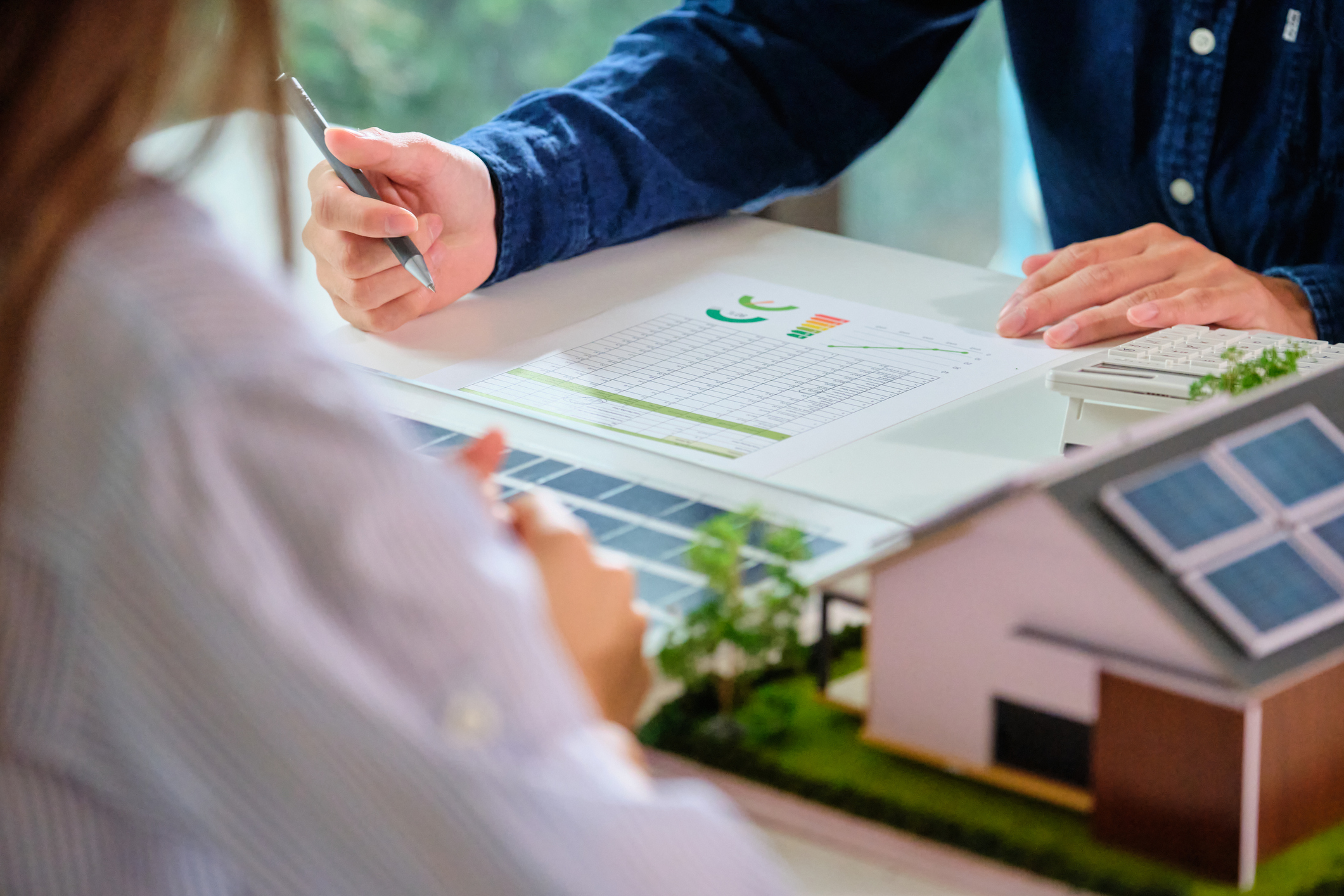 Nahaufnahme einer professionellen Beratung zur CO2-Kostenaufteilung Vermieter. Eine Hand zeigt auf Diagramme und Tabellen mit Energiedaten. Im Hintergrund sind ein Taschenrechner und ein Modellhaus mit Photovoltaikanlage zu sehen. Das Bild symbolisiert die wirtschaftliche Optimierung der CO2-Bilanz eines Mehrfamilienhauses durch PV-Sharing, um die Kostenlast bei einem CO2-Preis von 65 Euro zu senken.