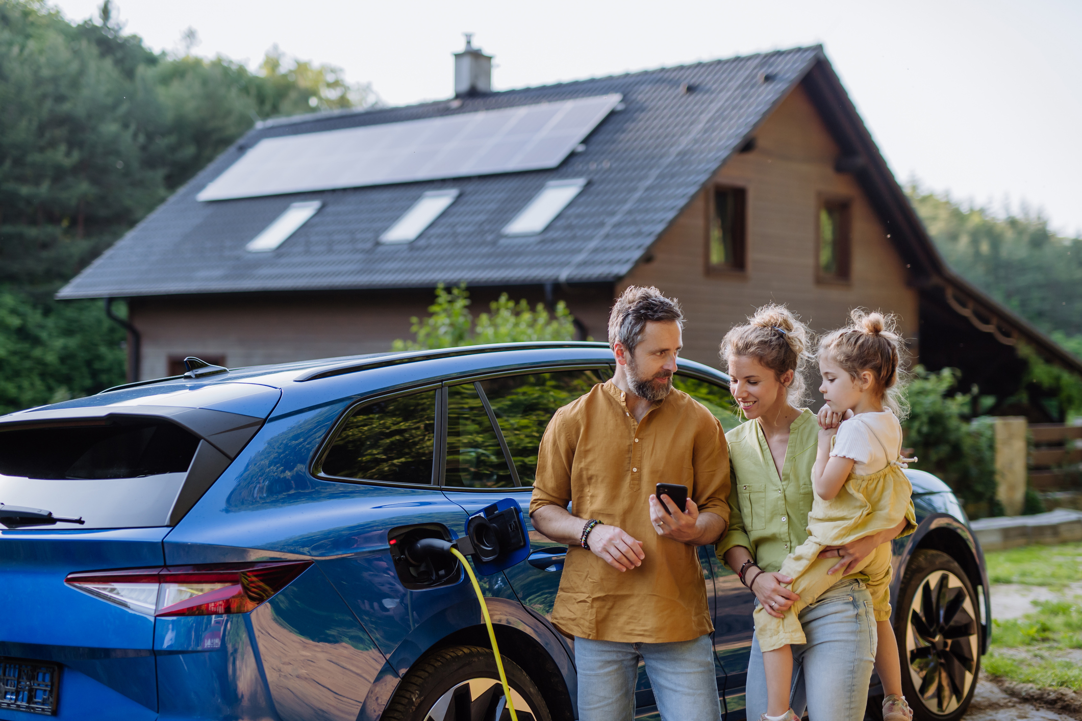 Eine Familie steht neben einem ladenden Elektroauto vor einem Wohngebäude mit Photovoltaikanlage zur Sektorenkopplung im Mehrfamilienhaus.